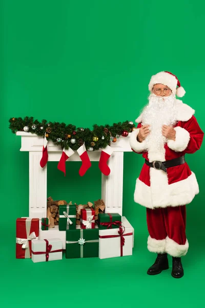 Barbe Père Noël claus debout près de cadeaux et cheminée décorée sur fond vert — Photo de stock