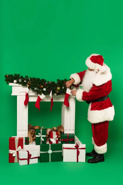 Santa Claude tenant bas de Noël près de cheminée décorée et présente sur fond vert — Photo de stock