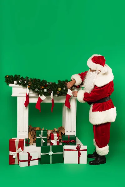 Santa Claus celebración de medias de Navidad cerca de la chimenea decorada y regalos sobre fondo verde - foto de stock