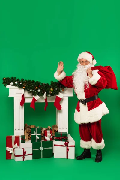 Santa Claus con saco saludando a la cámara cerca de la chimenea y regalos sobre fondo verde - foto de stock