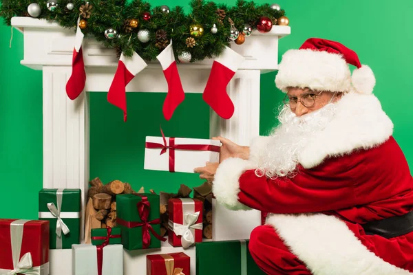 Santa claus holding present near fireplace with fir branches and baubles on green background — Stock Photo