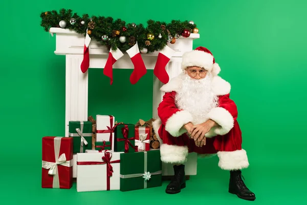 Санта-Клаус смотрит на камеру рядом с подарками и украшенный камин на зеленом фоне — стоковое фото