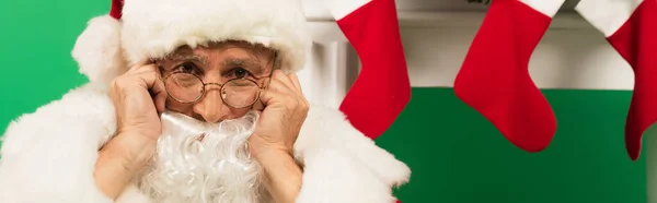 Sad santa claus looking at camera near christmas stockings on fireplace on green background, banner — Stock Photo
