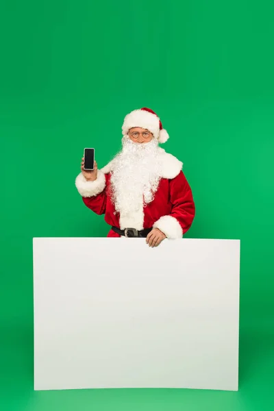 Weihnachtsmann hält Smartphone mit leerem Bildschirm und Plakat auf grünem Hintergrund — Stockfoto