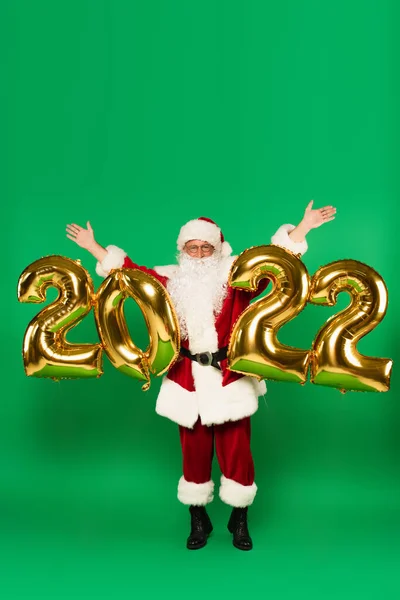 Santa claus raising hands near balloons in shape of 2022 on green background — Stock Photo