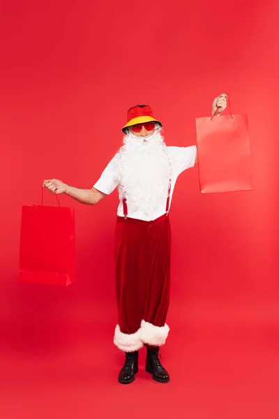 Санта-Клаус в сонцезахисних окулярах тримає сумки на червоному тлі — стокове фото