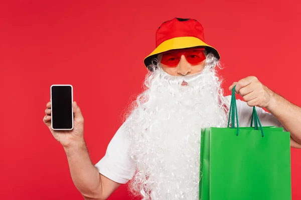 Santa Claus en gafas de sol con bolsas de compras y smartphone con pantalla en blanco aislada en rojo - foto de stock