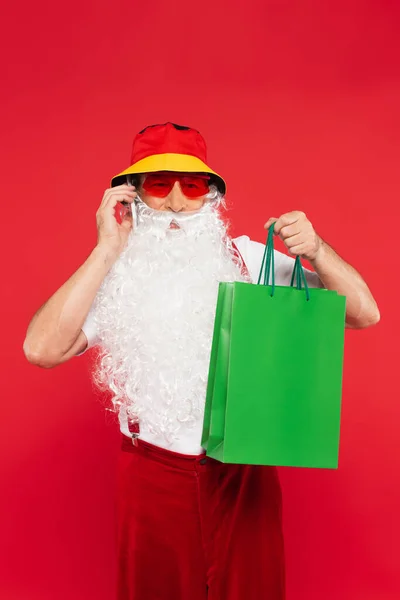 Santa Claude en panama et lunettes de soleil parlant sur smartphone et tenant des sacs à provisions isolés sur rouge — Photo de stock