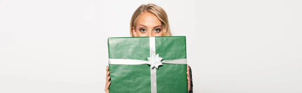 Mujer rubia oscureciendo la cara con caja de regalo verde y mirando a la cámara aislada en blanco, pancarta - foto de stock