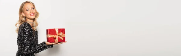 Gioiosa donna in camicetta rossa con paillettes in possesso di regalo e sorridente alla macchina fotografica su bianco, banner — Foto stock
