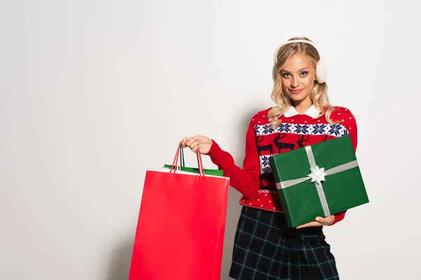 Mujer feliz en orejeras y suéter cálido posando con bolsas de compras y caja de regalo en blanco - foto de stock