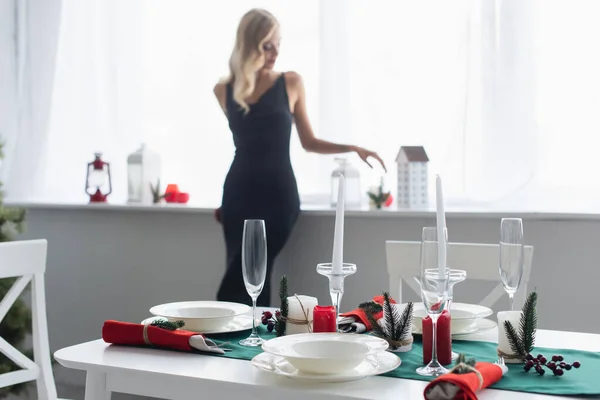 Messa a fuoco selettiva del tavolo servito per la cena di Natale vicino donna elegante in piedi dalla finestra su sfondo sfocato — Foto stock