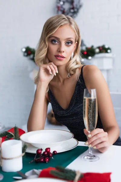 Hübsche und elegante Frau blickt in die Kamera, während sie am festlichen Tisch mit einem Glas Champagner sitzt — Stockfoto