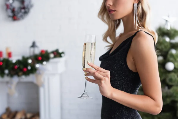 Vista parcial de la mujer elegante con copa de champán en la sala de estar con decoración de Navidad borrosa - foto de stock