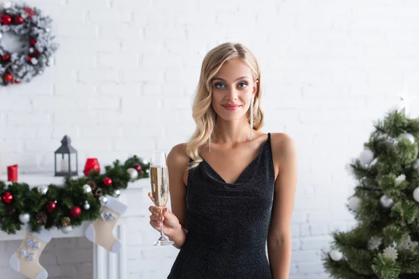 Elegante mujer rubia con copa de champán sonriendo a la cámara cerca borrosa árbol de Navidad y chimenea decorada - foto de stock