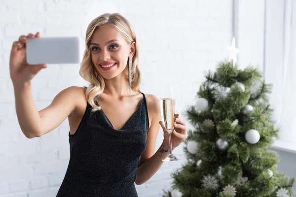 Donna allegra in abito elegante prendendo selfie con bicchiere di champagne vicino albero di Natale offuscata — Foto stock