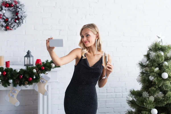 Feliz joven con copa de champán tomando selfie en el teléfono inteligente cerca del árbol de Navidad - foto de stock