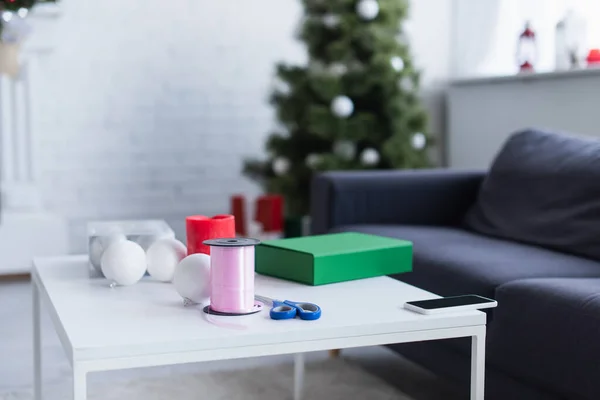 Tavolo con confezione regalo, bobina con nastro decorativo, palline e forbici in soggiorno con albero di Natale sfocato — Foto stock