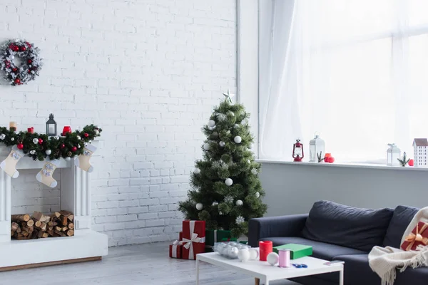 Espaçosa sala de estar com árvore de natal, lareira decorada, sofá e mesa com bolas de Natal e bobina de fita decorativa — Fotografia de Stock