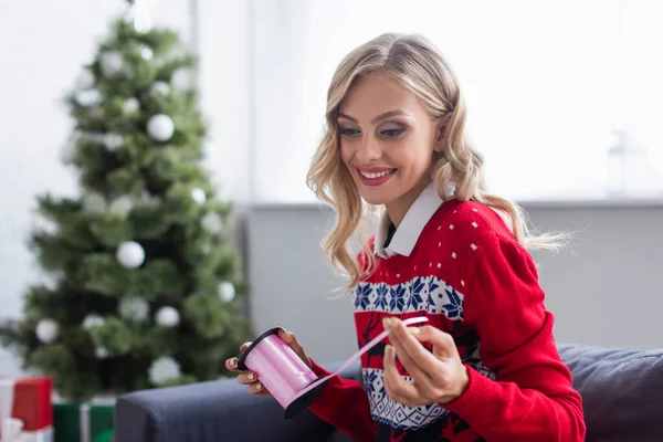 Femme souriante en pull tendance tenant bobine avec ruban décoratif près de l'arbre de Noël flou — Photo de stock