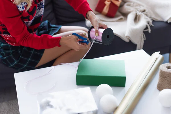 Vista recortada de la mujer cortando cinta decorativa cerca de la caja de regalo, bobina con cordel, papel de envolver y bolas de Navidad - foto de stock