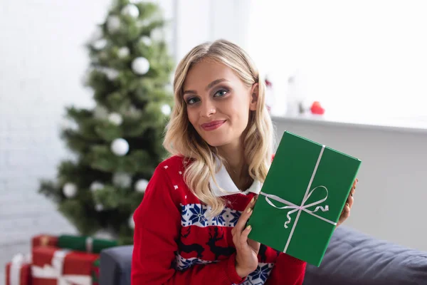 Весела блондинка в теплому светрі тримає зелену подарункову коробку біля ялинки на розмитому фоні — стокове фото