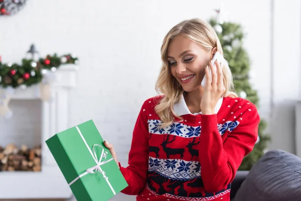 Mulher sorridente em camisola elegante com ornamento segurando presente de Natal enquanto fala no smartphone — Fotografia de Stock