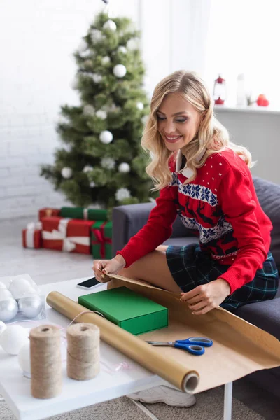 Happy woman in warm sweater packing gift box into wrapping paper near presents under blurred christmas tree — Stock Photo