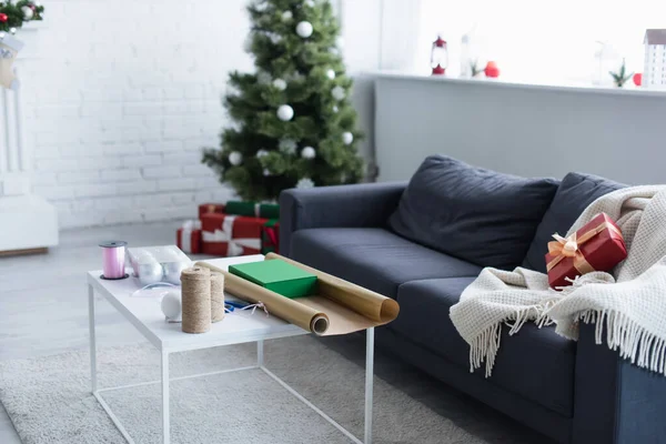 Sofá com cobertor quente e caixa de presente perto da mesa com fio, fita decorativa e papel de embrulho perto da árvore de Natal borrada — Fotografia de Stock