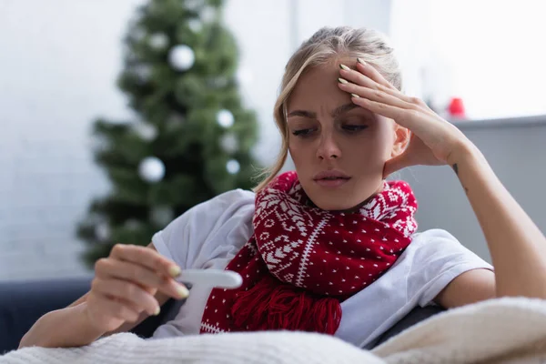 Donna preoccupata in sciarpa calda guardando termometro vicino albero di Natale su sfondo sfocato — Foto stock