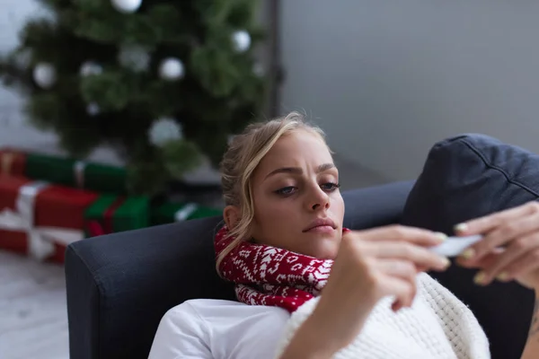 Donna scontenta e malata sul divano guardando il termometro vicino all'albero di Natale su sfondo sfocato — Foto stock