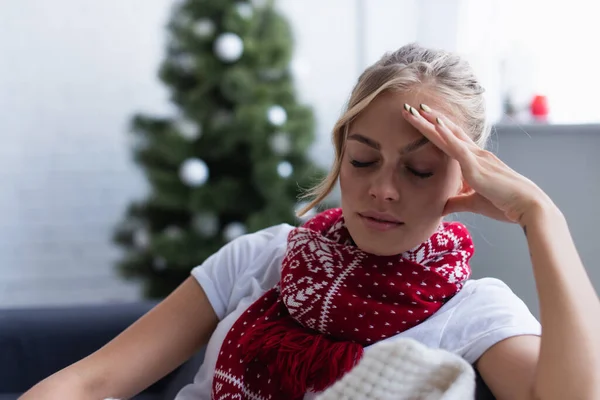 Femme malade en écharpe chaude assise sur le canapé avec les yeux fermés près de l'arbre de Noël sur fond flou — Photo de stock