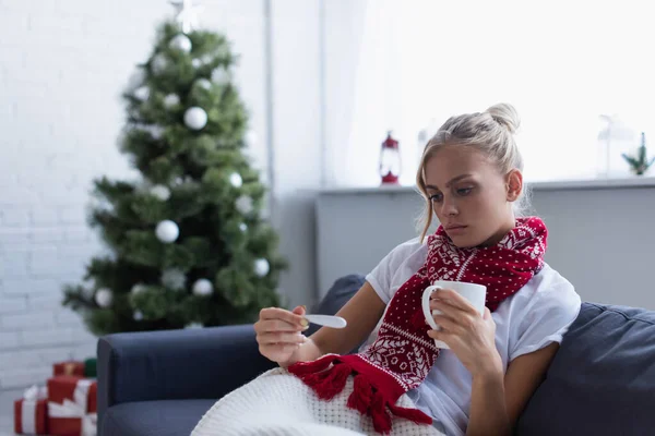 Upset woman with warm drink and thermometer near christmas tree on blurred background — Stock Photo