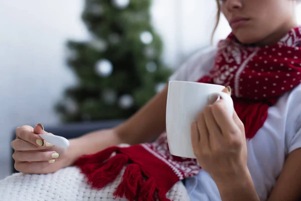 Vista cortada de mulher doente com termômetro e xícara de bebida quente perto da árvore de natal no fundo borrado — Fotografia de Stock