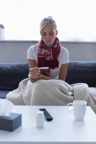Upset woman in warm scarf looking at thermometer near blurred pills container and warm drink — Stock Photo