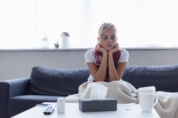 Traurige und kranke Frau in warmem Schal sitzt auf Sofa unter Decke neben Papierservietten und Tablettenbehälter — Stockfoto