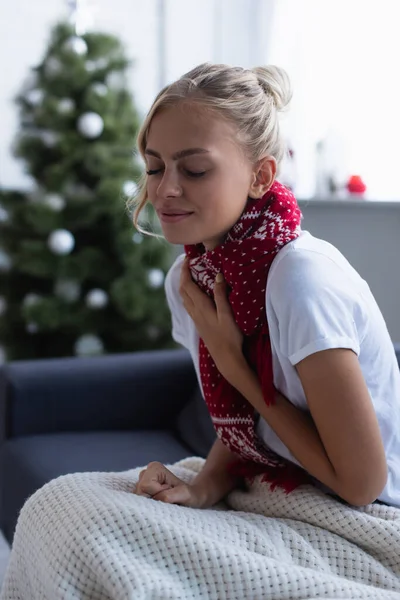 Femme malade dans l'écharpe chaude toussant sur le canapé avec les yeux fermés près de l'arbre de Noël flou — Photo de stock