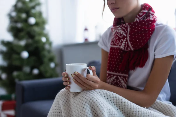 Cropped view of ill woman in scarf sitting under blanket with cup of warm drink near blurred christmas tree — Stock Photo