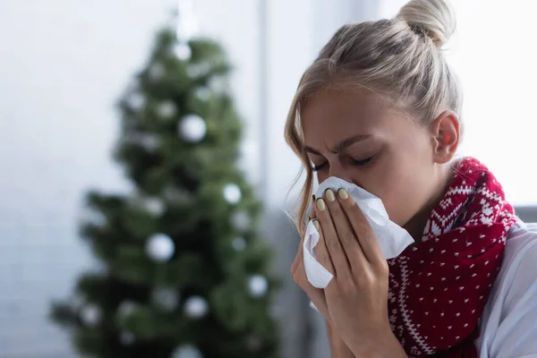 Femme malade éternuer dans une serviette en papier près de l'arbre de Noël sur fond flou — Photo de stock