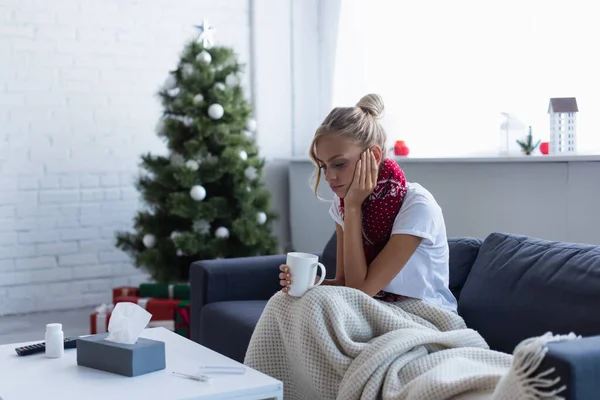 Femme malade de mauvaise humeur assis avec une tasse de boisson chaude près de l'arbre de Noël sur fond flou — Photo de stock