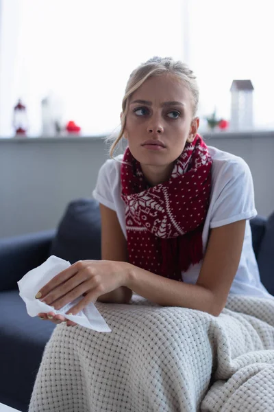 Kranke und nachdenkliche Frau in warmem Schal sitzt auf Sofa mit Papierserviette — Stockfoto