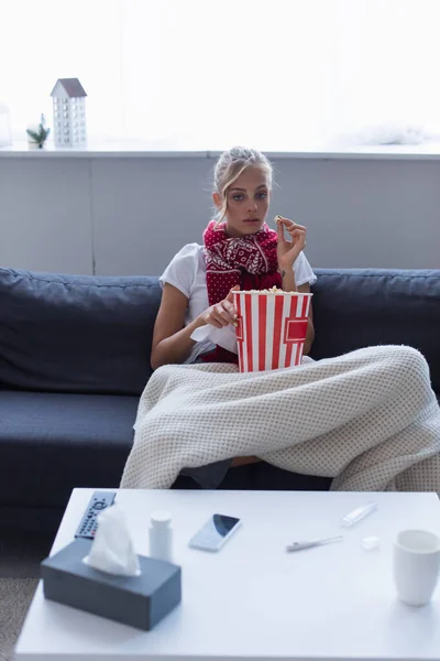 Mujer enferma viendo la televisión con cubo de palomitas de maíz cerca del termómetro y píldoras contenedor en la mesa - foto de stock