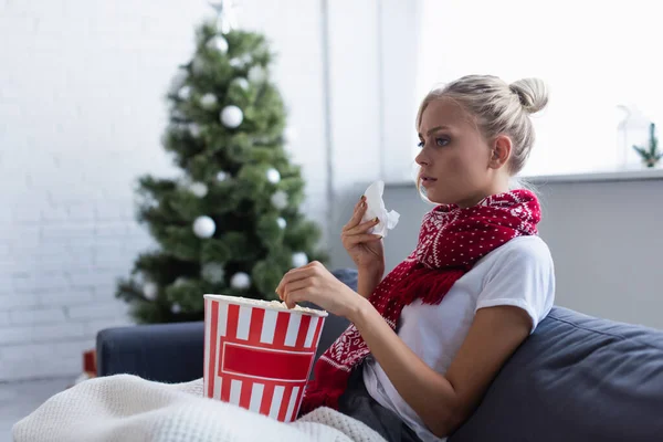 Donna malata con tovagliolo di carta e secchio di popcorn guardando la tv sul divano vicino all'albero di Natale offuscato — Foto stock