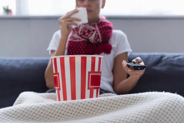 Cropped view of sick woman with remote controller and bucket of popcorn watching tv on blurred background — Stock Photo