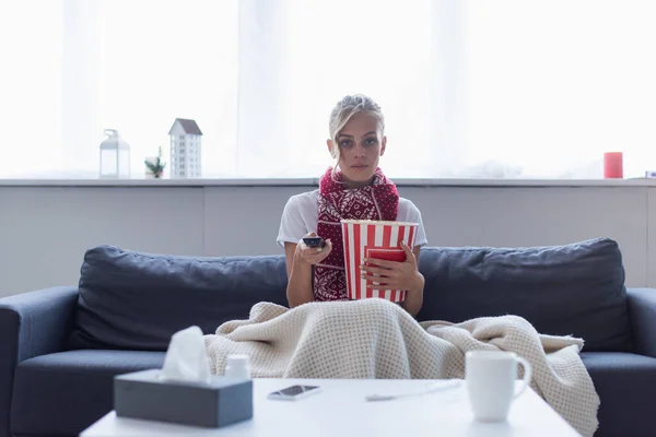 Mujer enferma con mando a distancia y cubo de palomitas de maíz viendo televisión cerca de servilletas de papel borroso y taza de bebida caliente - foto de stock