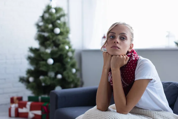 Donna malata e triste guardando altrove mentre seduto sul divano vicino all'albero di Natale offuscato — Foto stock