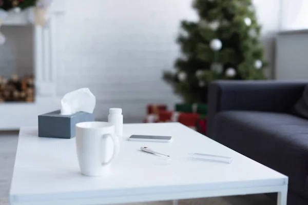 Table avec serviettes en papier, récipient à pilules, thermomètre électronique et smartphone près de l'arbre de Noël flou — Photo de stock