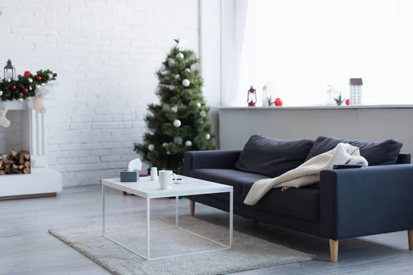 Couverture chaude sur le canapé près de la table avec récipient de pilules, tasse de boisson chaude et serviettes en papier dans le salon avec arbre de Noël décoré — Photo de stock