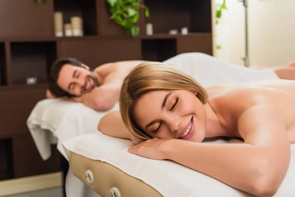 Femme souriante avec les yeux fermés couché sur la table de massage près du petit ami dans le centre de spa — Photo de stock