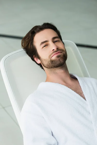 Hombre joven en albornoz blanco mirando a la cámara en la silla de cubierta en el centro de spa - foto de stock